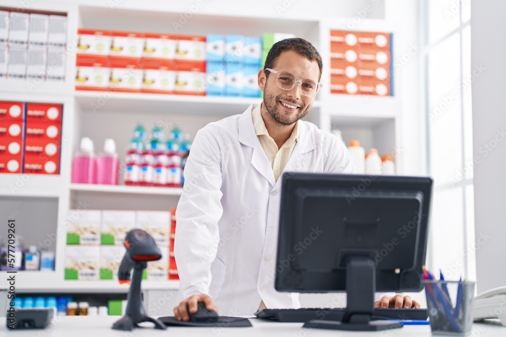 Young man pharmacist smiling confident using computer at pharmacy