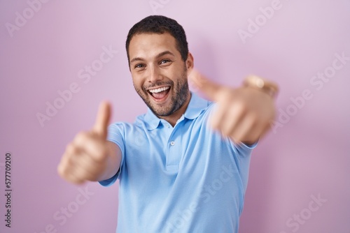 Hispanic man standing over pink background approving doing positive gesture with hand, thumbs up smiling and happy for success. winner gesture.