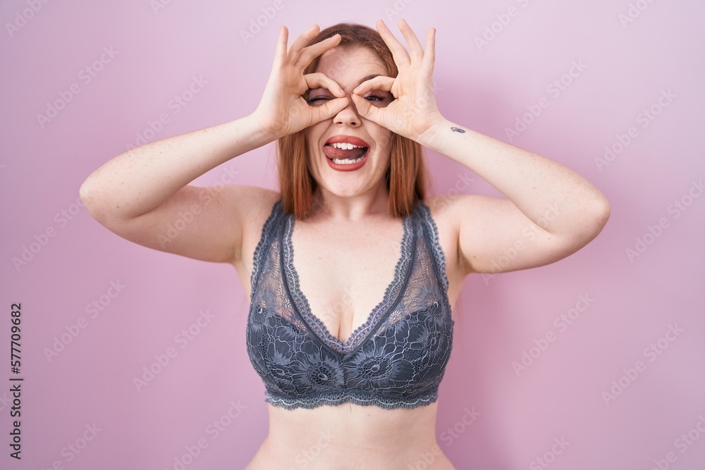 Redhead woman wearing lingerie over pink background doing ok gesture like binoculars sticking tongue out, eyes looking through fingers. crazy expression. Redhead woman wearing lingerie over pink background doing ok gesture like binoculars sticking tongue out, eyes looking through fingers. crazy expression.