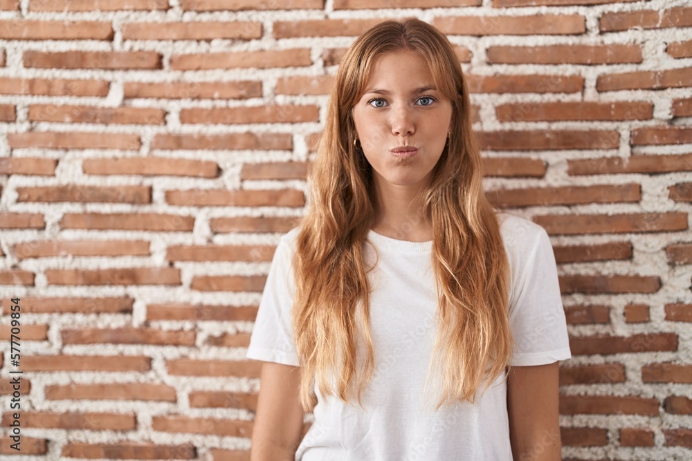Young caucasian woman standing over bricks wall puffing cheeks with funny face. mouth inflated with air, crazy expression.