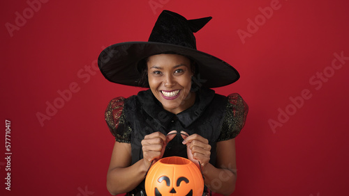 African american woman wearing witch costume holding halloween pumpkin basket over isolated red background