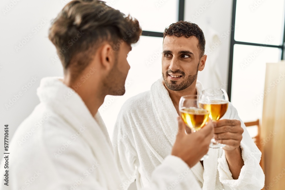 Two hispanic men couple toasting with champagne sitting on massage table at beauty center Two hispanic men couple toasting with champagne sitting on massage table at beauty center
