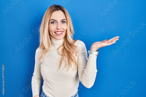Young caucasian woman standing over blue background smiling cheerful presenting and pointing with palm of hand looking at the camera.