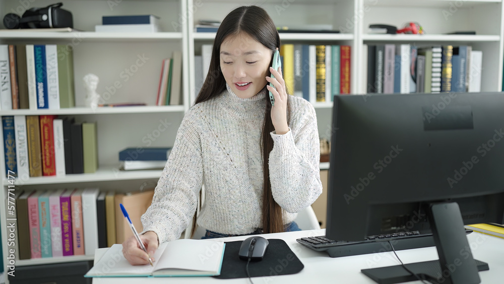 Young chinese woman student talking on smartphone writing on notebook at library university Young chinese woman student talking on smartphone writing on notebook at library university