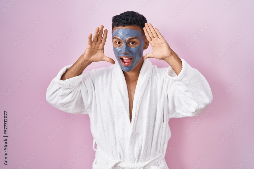 Young hispanic man wearing beauty face mask and bath robe smiling cheerful playing peek a boo with hands showing face. surprised and exited Young hispanic man wearing beauty face mask and bath robe smiling cheerful playing peek a boo with hands showing face. surprised and exited