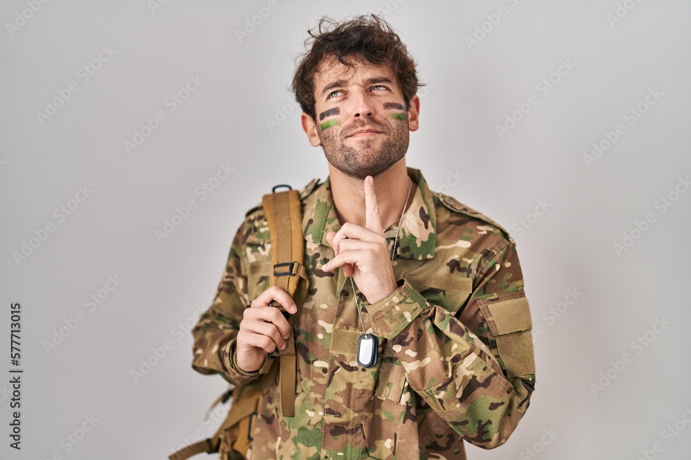 Hispanic young man wearing camouflage army uniform thinking concentrated about doubt with finger on chin and looking up wondering