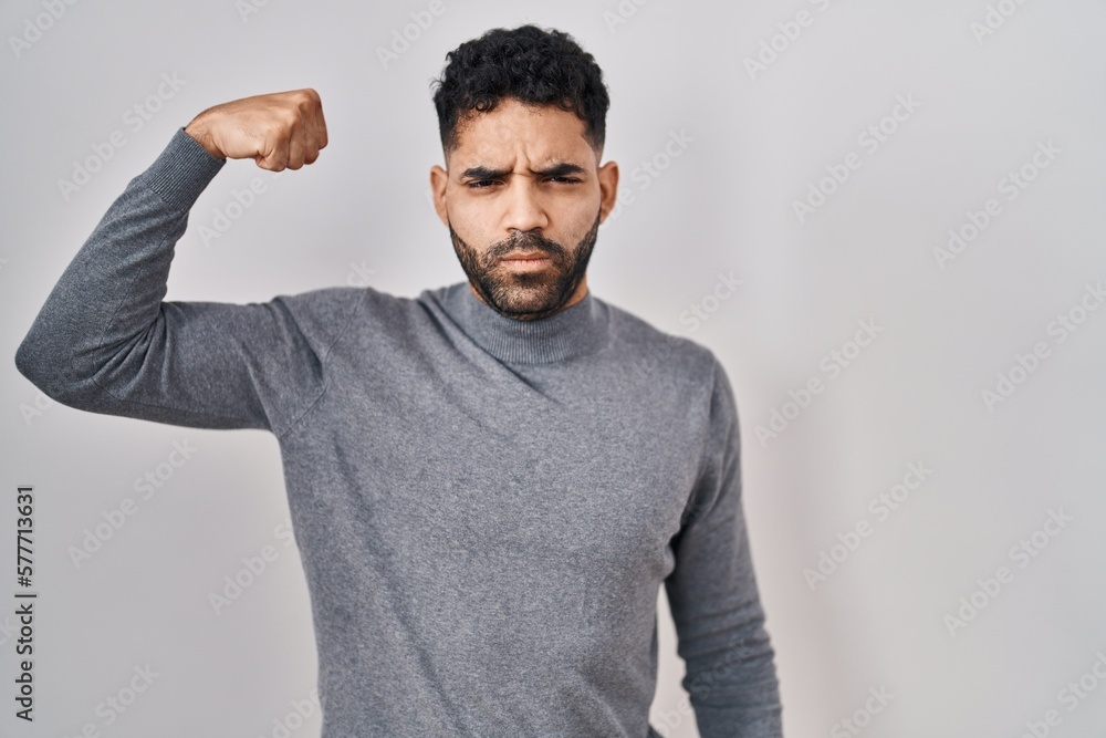 Hispanic man with beard standing over white background strong person showing arm muscle, confident and proud of power Hispanic man with beard standing over white background strong person showing arm muscle, confident and proud of power