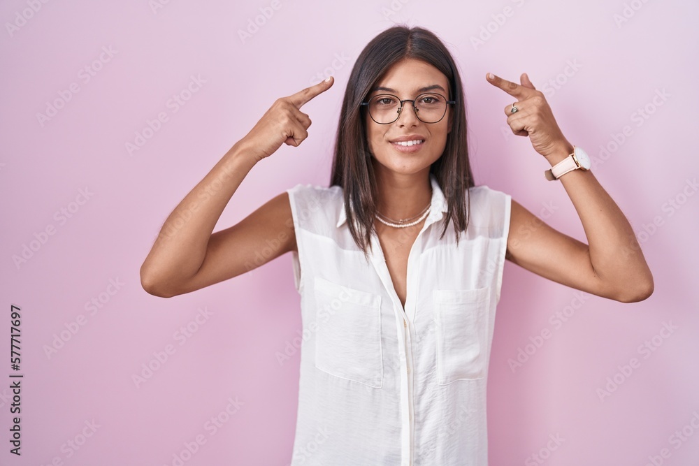 Brunette young woman standing over pink background wearing glasses smiling pointing to head with both hands finger, great idea or thought, good memory Brunette young woman standing over pink background wearing glasses smiling pointing to head with both hands finger, great idea or thought, good memory