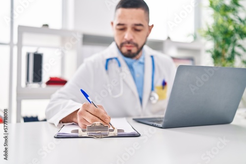 Young latin man doctor using laptop writing medical report at clinic