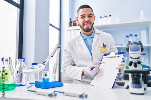 Hispanic man working at scientist laboratory holding blank clipboard sticking tongue out happy with funny expression.