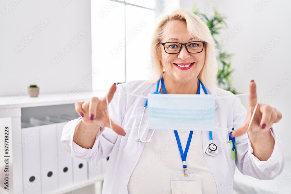 Middle age blonde woman wearing doctor uniform holding medical mask at clinic Middle age blonde woman wearing doctor uniform holding medical mask at clinic