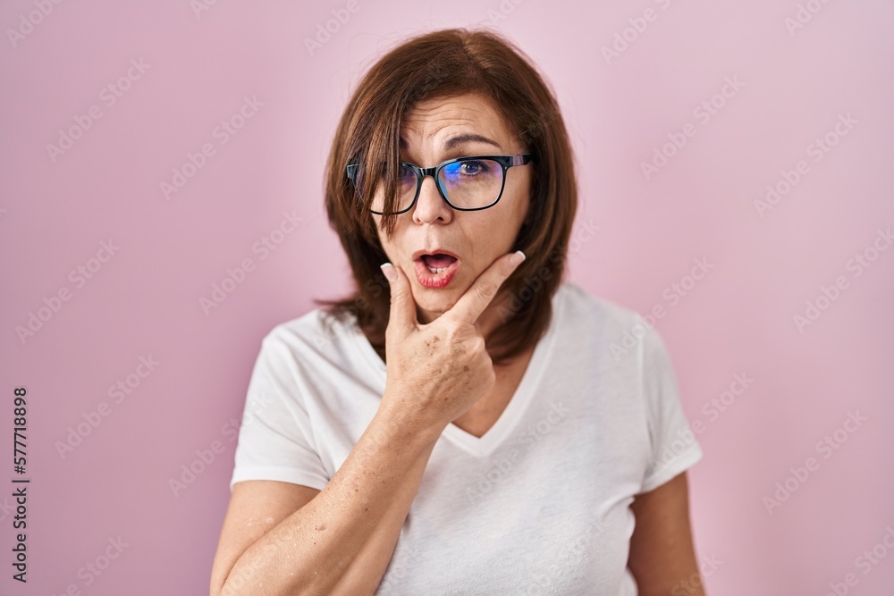 Middle age hispanic woman standing over pink background looking fascinated with disbelief, surprise and amazed expression with hands on chin Middle age hispanic woman standing over pink background looking fascinated with disbelief, surprise and amazed expression with hands on chin