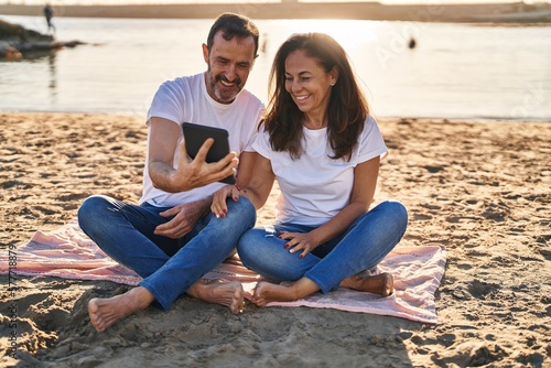 Middle age man and woman couple using touchpad sitting on sand at seaside