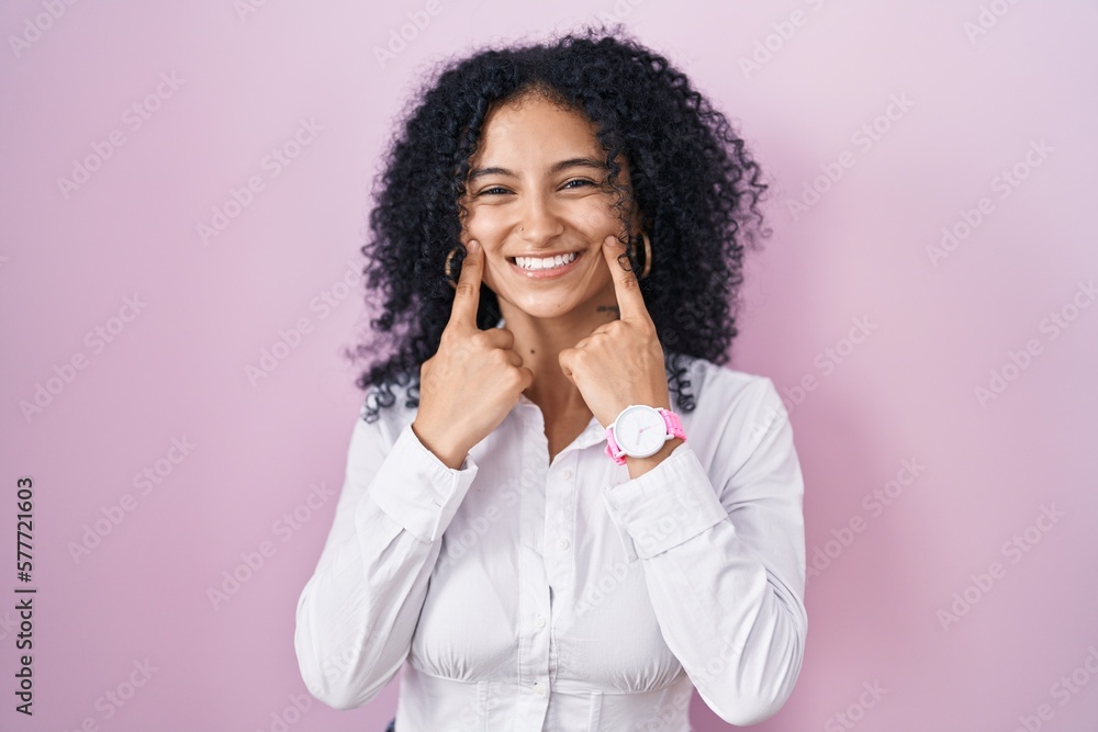 Hispanic woman with curly hair standing over pink background smiling with open mouth, fingers pointing and forcing cheerful smile Hispanic woman with curly hair standing over pink background smiling with open mouth, fingers pointing and forcing cheerful smile