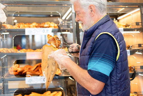 Smiling senior man buying freshly baked croissants in supermarket shopping for groceries paying attention to cost and quality - concept of consumerism, price increase, inflation
