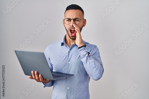 Young hispanic man working using computer laptop shouting and screaming loud to side with hand on mouth. communication concept.