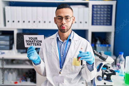 Young hispanic man working at scientist laboratory holding your donation matters holding blood sample puffing cheeks with funny face. mouth inflated with air, catching air.