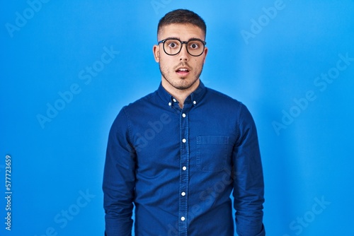 Young hispanic man wearing glasses over blue background afraid and shocked with surprise expression  fear and excited face.