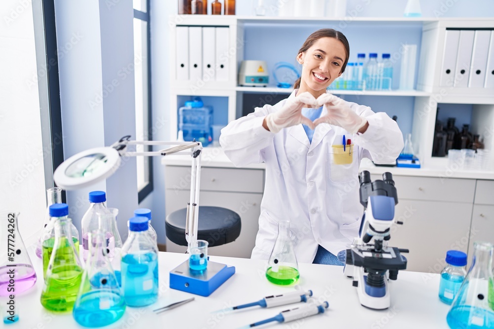 Young brunette woman working at scientist laboratory smiling in love showing heart symbol and shape with hands. romantic concept. Young brunette woman working at scientist laboratory smiling in love showing heart symbol and shape with hands. romantic concept.