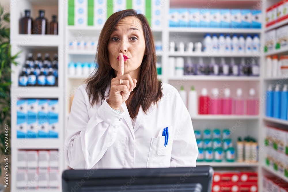 Middle age brunette woman working at pharmacy drugstore asking to be quiet with finger on lips. silence and secret concept.