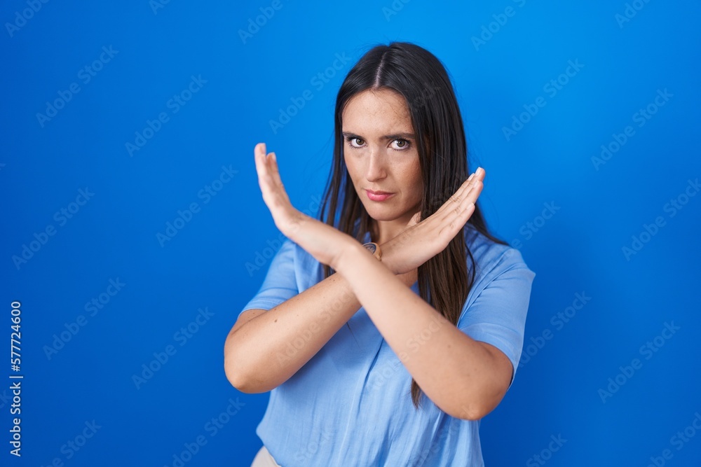 Fototapeta premium Young brunette woman standing over blue background rejection expression crossing arms doing negative sign, angry face