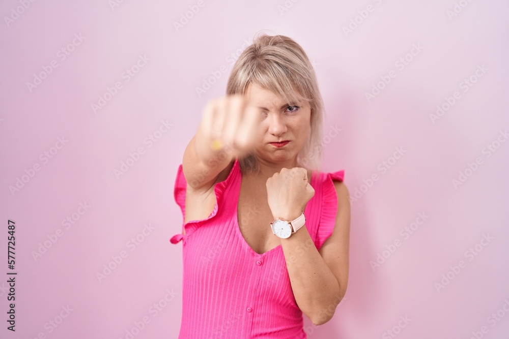 Young caucasian woman standing over pink background punching fist to fight, aggressive and angry attack, threat and violence Young caucasian woman standing over pink background punching fist to fight, aggressive and angry attack, threat and violence