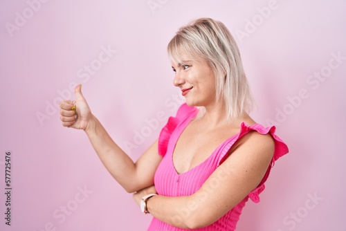 Young caucasian woman standing over pink background looking proud, smiling doing thumbs up gesture to the side © Krakenimages.com