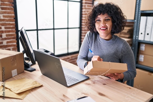 Young beautiful hispanic woman ecommerce business worker writing on package at office