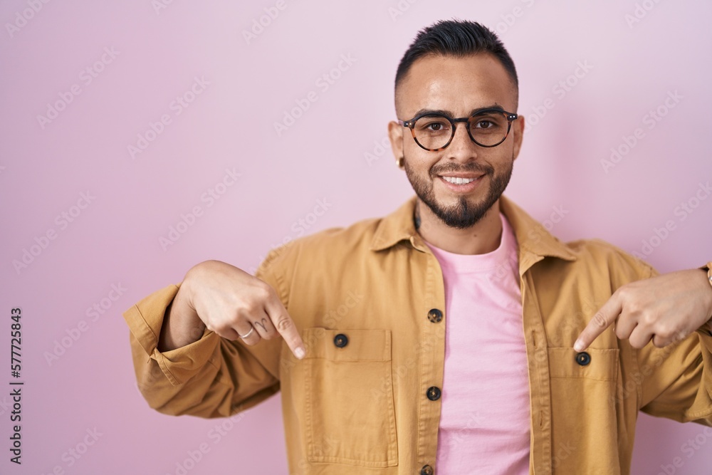 Young hispanic man standing over pink background looking confident with smile on face, pointing oneself with fingers proud and happy.