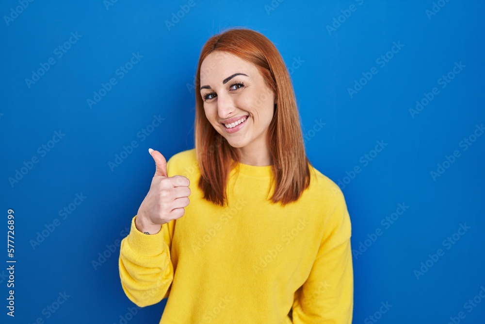 Young woman standing over blue background doing happy thumbs up gesture with hand. approving expression looking at the camera showing success. Young woman standing over blue background doing happy thumbs up gesture with hand. approving expression looking at the camera showing success.