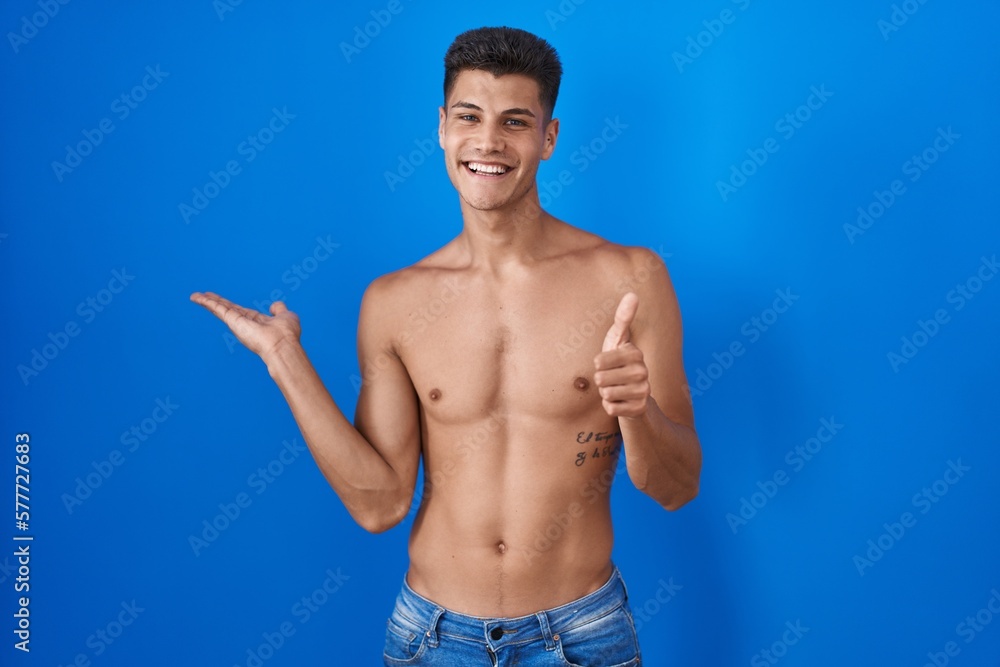 Young hispanic man standing shirtless over blue background showing palm hand and doing ok gesture with thumbs up, smiling happy and cheerful