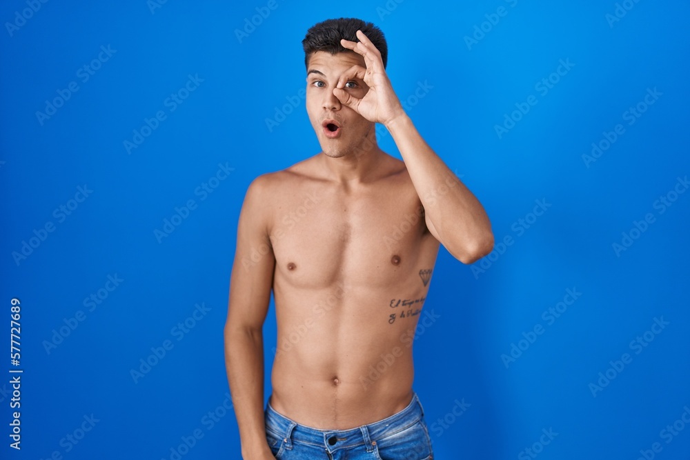 Young hispanic man standing shirtless over blue background doing ok gesture shocked with surprised face, eye looking through fingers. unbelieving expression.