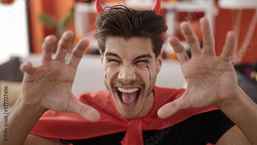 Young hispanic man wearing devil costume doing scare gesture at home