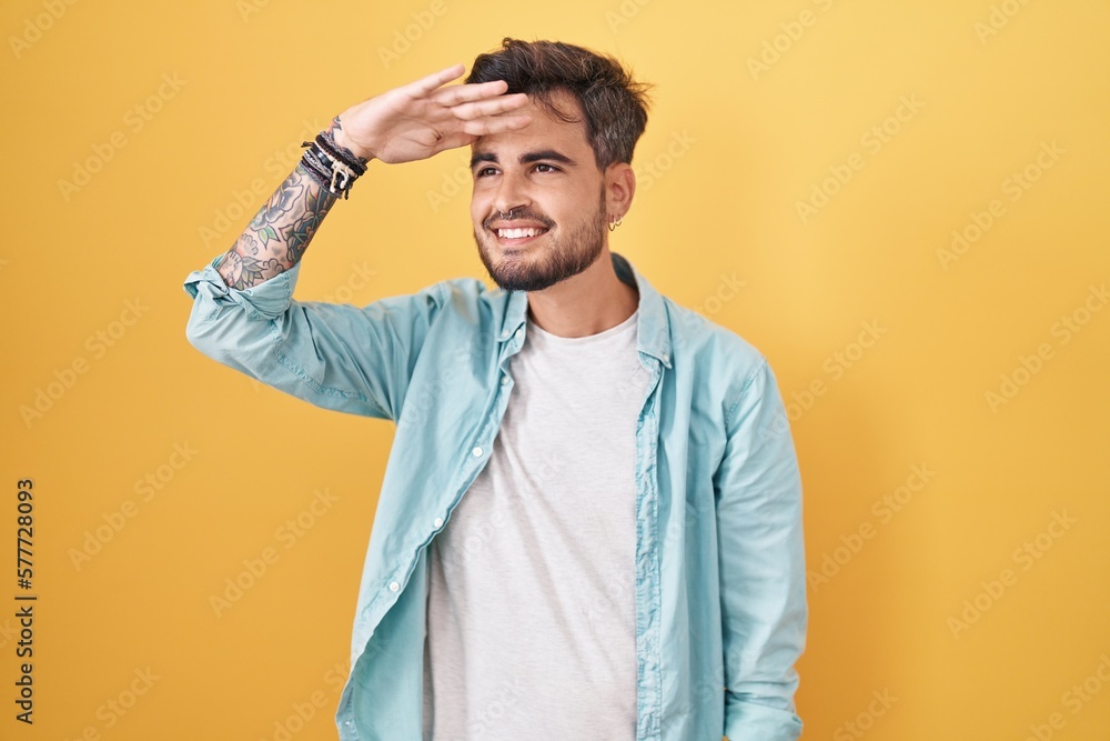Young hispanic man with tattoos standing over yellow background very happy and smiling looking far away with hand over head. searching concept. Young hispanic man with tattoos standing over yellow background very happy and smiling looking far away with hand over head. searching concept.