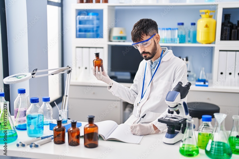 Young hispanic man scientist writing on notebook holding bottle at laboratory Young hispanic man scientist writing on notebook holding bottle at laboratory