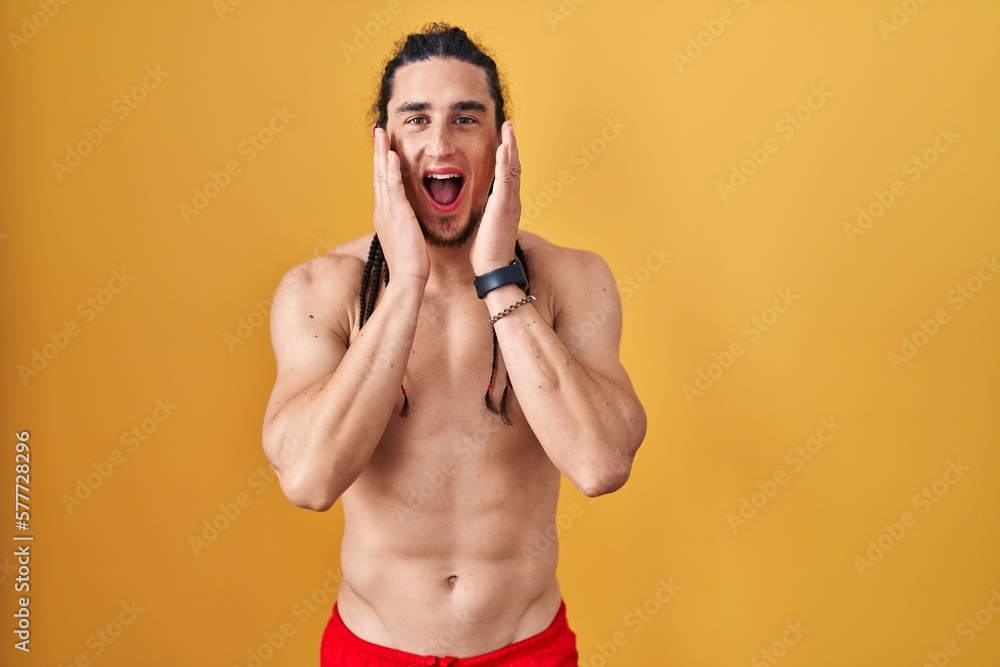 Hispanic man with long hair standing shirtless over yellow background afraid and shocked, surprise and amazed expression with hands on face Hispanic man with long hair standing shirtless over yellow background afraid and shocked, surprise and amazed expression with hands on face