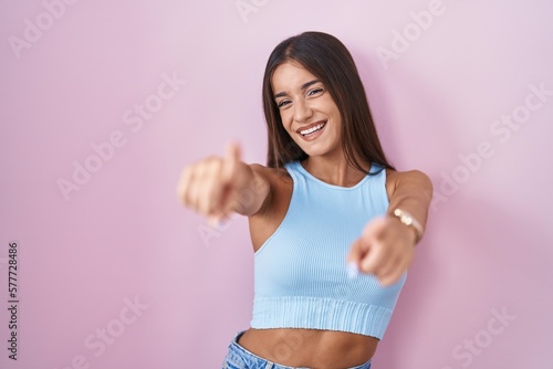 Young brunette woman standing over pink background pointing to you and the camera with fingers, smiling positive and cheerful © Krakenimages.com