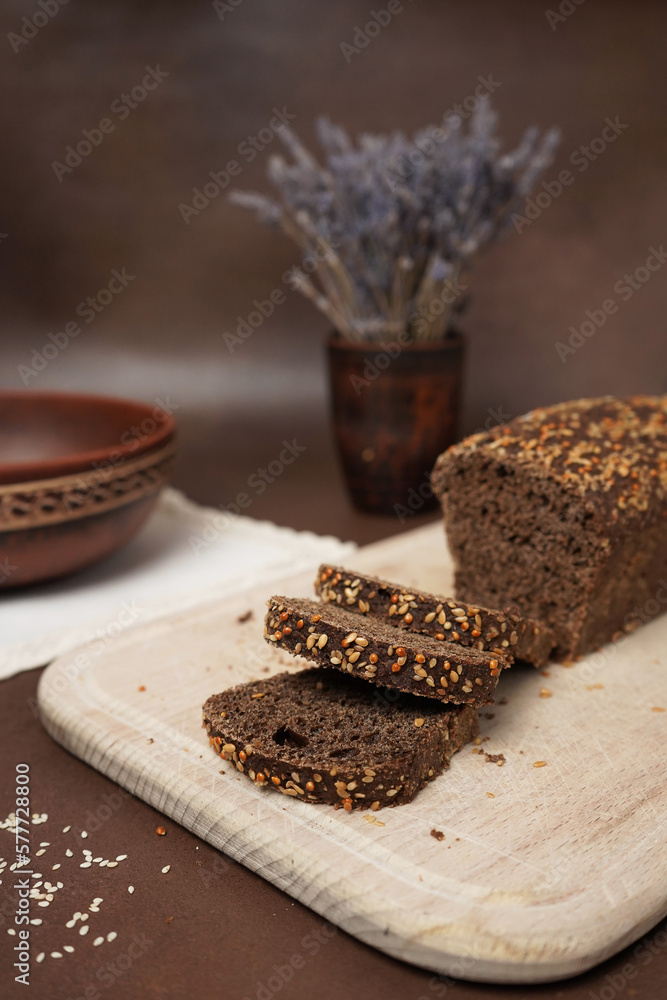 Black Bread with cereals Sliced on a Wooden cutting Close up Board against a brown background. Next to the bread is a Clay Plate, fork, spoon and white cotton tablecloth