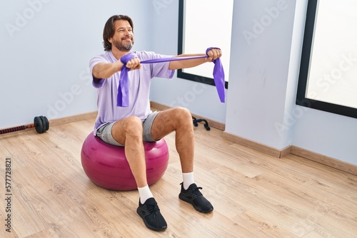 Middle age man training with elastic band sitting on fit ball at sport center