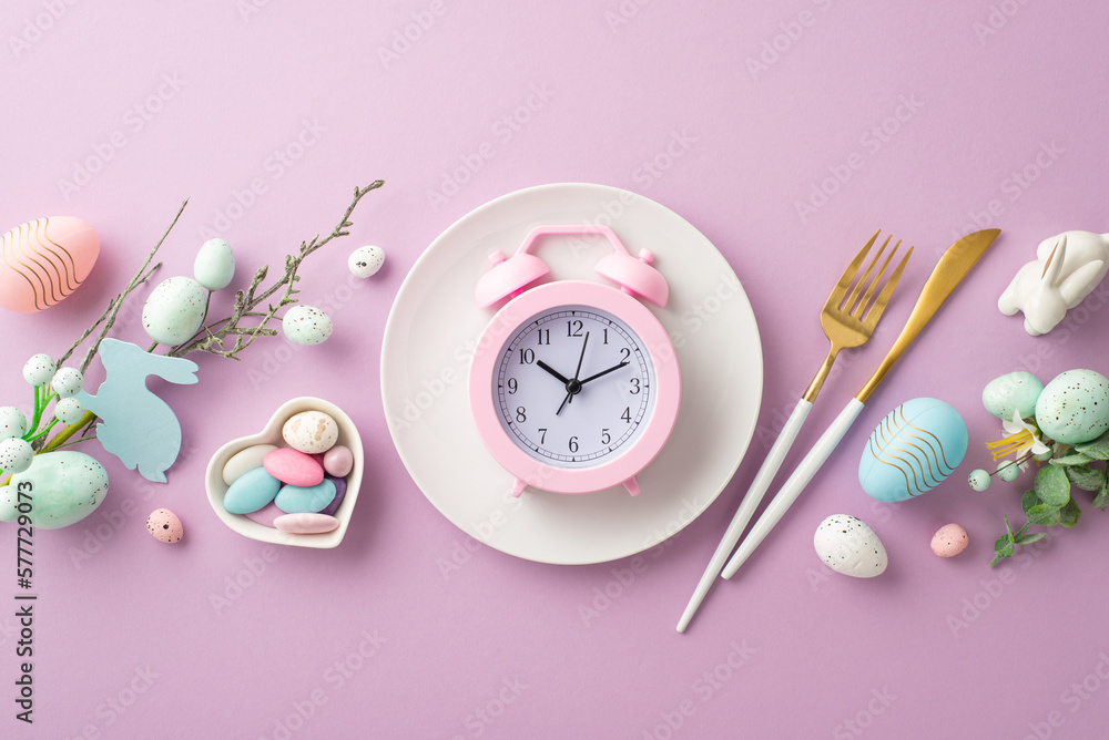 Easter celebration concept. Top view photo of pink alarm clock in plate fork knife floral decor pink and blue eggs saucer with candies and ceramic bunny on isolated pastel violet background Easter celebration concept. Top view photo of pink alarm clock in plate fork knife floral decor pink and blue eggs saucer with candies and ceramic bunny on isolated pastel violet background