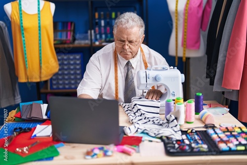 Middle age grey-haired man tailor using sewing machine and laptop at tailor shop