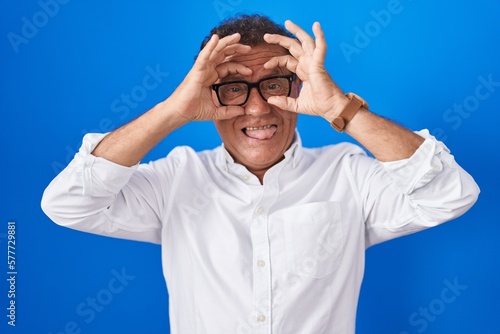 Middle age hispanic man standing over blue background doing ok gesture like binoculars sticking tongue out, eyes looking through fingers. crazy expression.