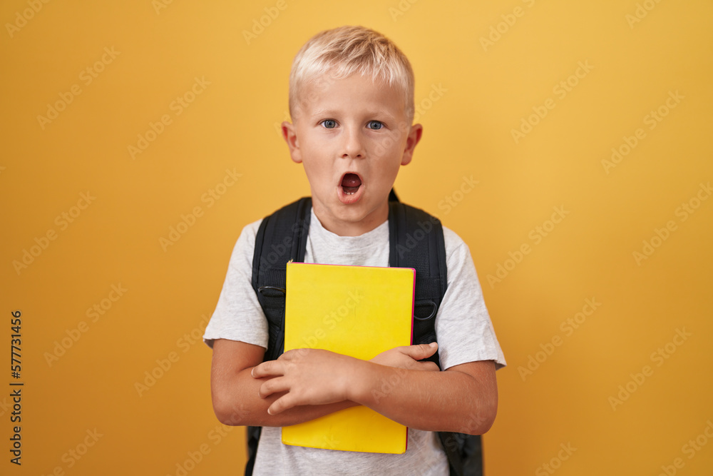 Little caucasian boy wearing student backpack and holding book scared and amazed with open mouth for surprise, disbelief face Little caucasian boy wearing student backpack and holding book scared and amazed with open mouth for surprise, disbelief face