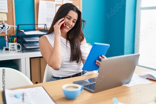 Young beautiful hispanic woman business worker talking on smartphone using touchpad at office