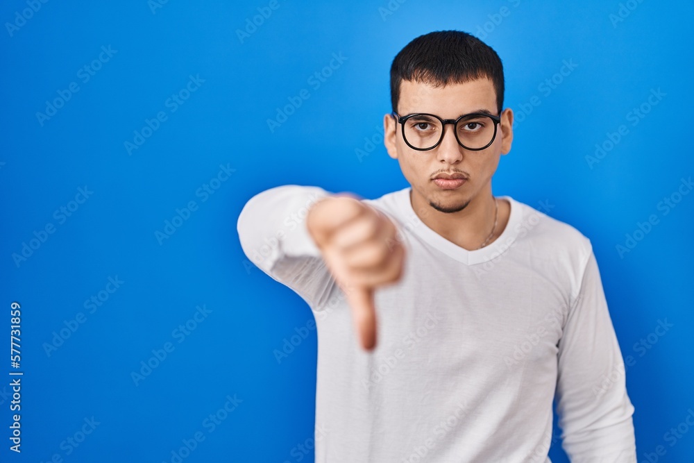 Young arab man wearing casual white shirt and glasses looking unhappy and angry showing rejection and negative with thumbs down gesture. bad expression. Young arab man wearing casual white shirt and glasses looking unhappy and angry showing rejection and negative with thumbs down gesture. bad expression.