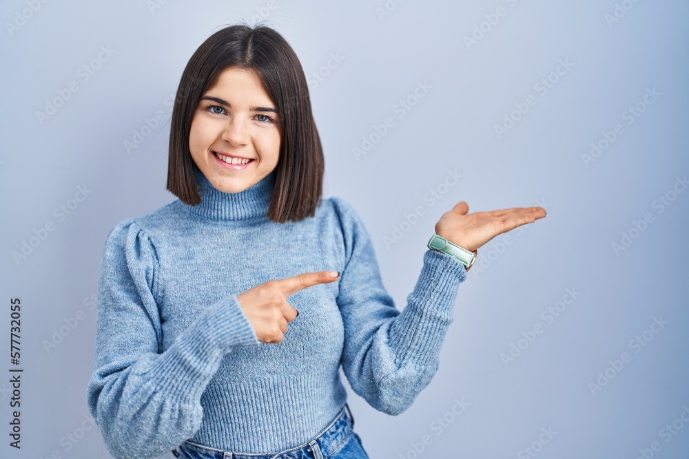Young hispanic woman standing over blue background amazed and smiling to the camera while presenting with hand and pointing with finger. Young hispanic woman standing over blue background amazed and smiling to the camera while presenting with hand and pointing with finger.