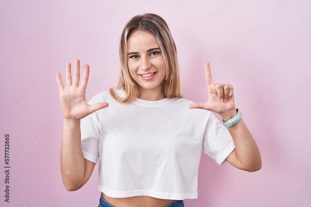 Young blonde woman standing over pink background showing and pointing up with fingers number seven while smiling confident and happy. Young blonde woman standing over pink background showing and pointing up with fingers number seven while smiling confident and happy.