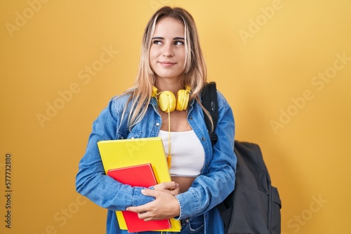Young blonde woman wearing student backpack and holding books smiling looking to the side and staring away thinking.