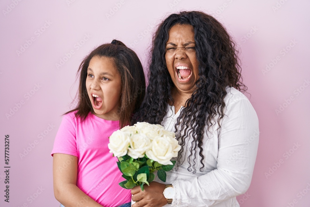 Mother and young daughter holding bouquet of white flowers angry and mad screaming frustrated and furious, shouting with anger. rage and aggressive concept. Mother and young daughter holding bouquet of white flowers angry and mad screaming frustrated and furious, shouting with anger. rage and aggressive concept.