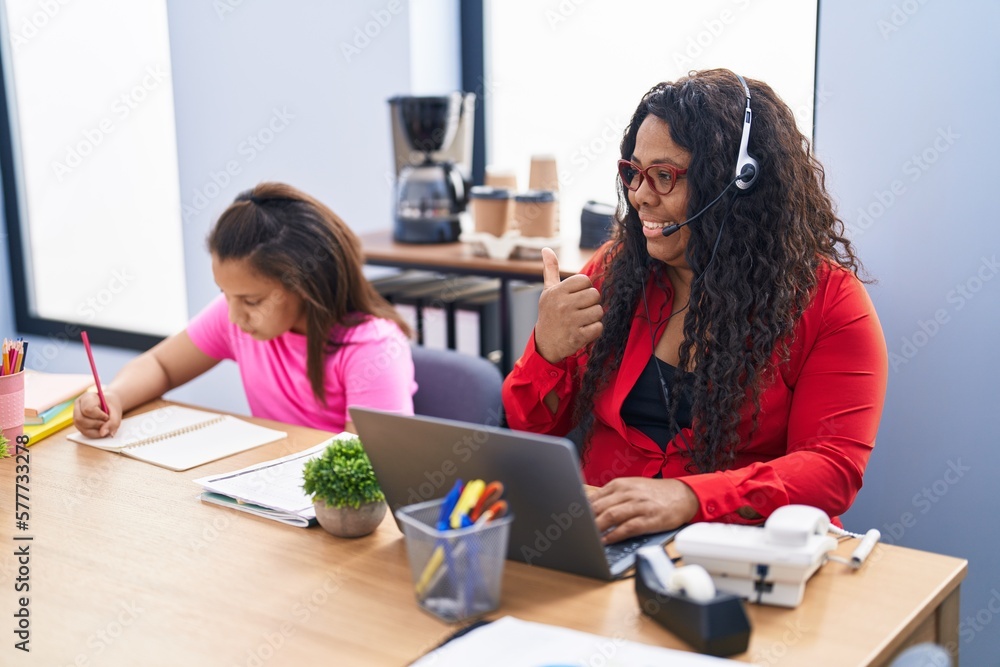 Mother and young daughter at the office working and doing homework pointing thumb up to the side smiling happy with open mouth Mother and young daughter at the office working and doing homework pointing thumb up to the side smiling happy with open mouth
